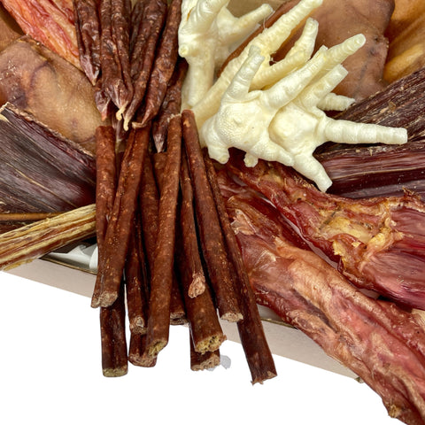 A detailed overhead image highlighting the texture of sausages, biltong, and chicken feet arranged within the Puppy’s First Feast box.